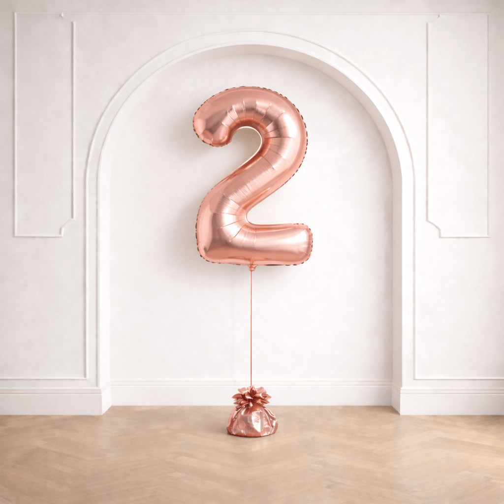 A rose gold number 2 foil balloon on a balloon weight in front of a white arched wall